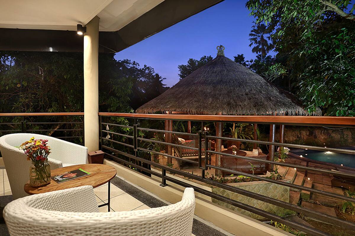 A balcony with wicker furniture, a thatched-roof structure, and a pool in the background.