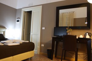 Una camera d'albergo con un letto, una scrivania con un computer, una sedia, un cestino e uno specchio a parete.