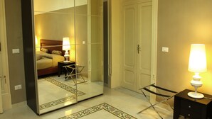 A hotel room with a large mirror, a bed with a headboard, a bedside table with a lamp, a small desk with a chair, and a wardrobe. - Abatellis Luxury (Palermo)