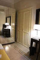Una camera d'albergo con un letto, una scrivania con una lampada, una sedia, un cestino e un armadio.