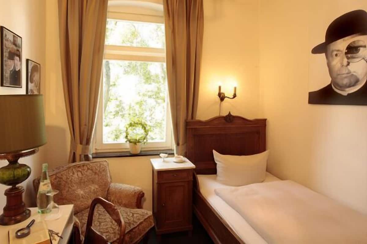 Une chambre d’hôtel comprenant un lit, un bureau avec une lampe, une chaise, une petite table avec un globe terrestre et une loupe, une table de chevet avec une lampe, et une fenêtre avec des rideaux.