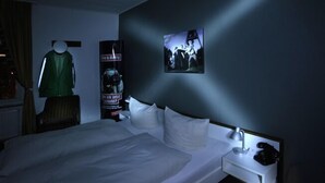 Habitación de hotel con una cama, una mesita de noche, una lámpara, un cartel y un maniquí vestido de verde.