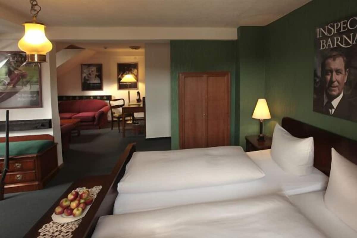Une chambre d’hôtel avec un grand lit, un bureau, une chaise et une lampe. Il y a une affiche accrochée au mur et une porte en bois.