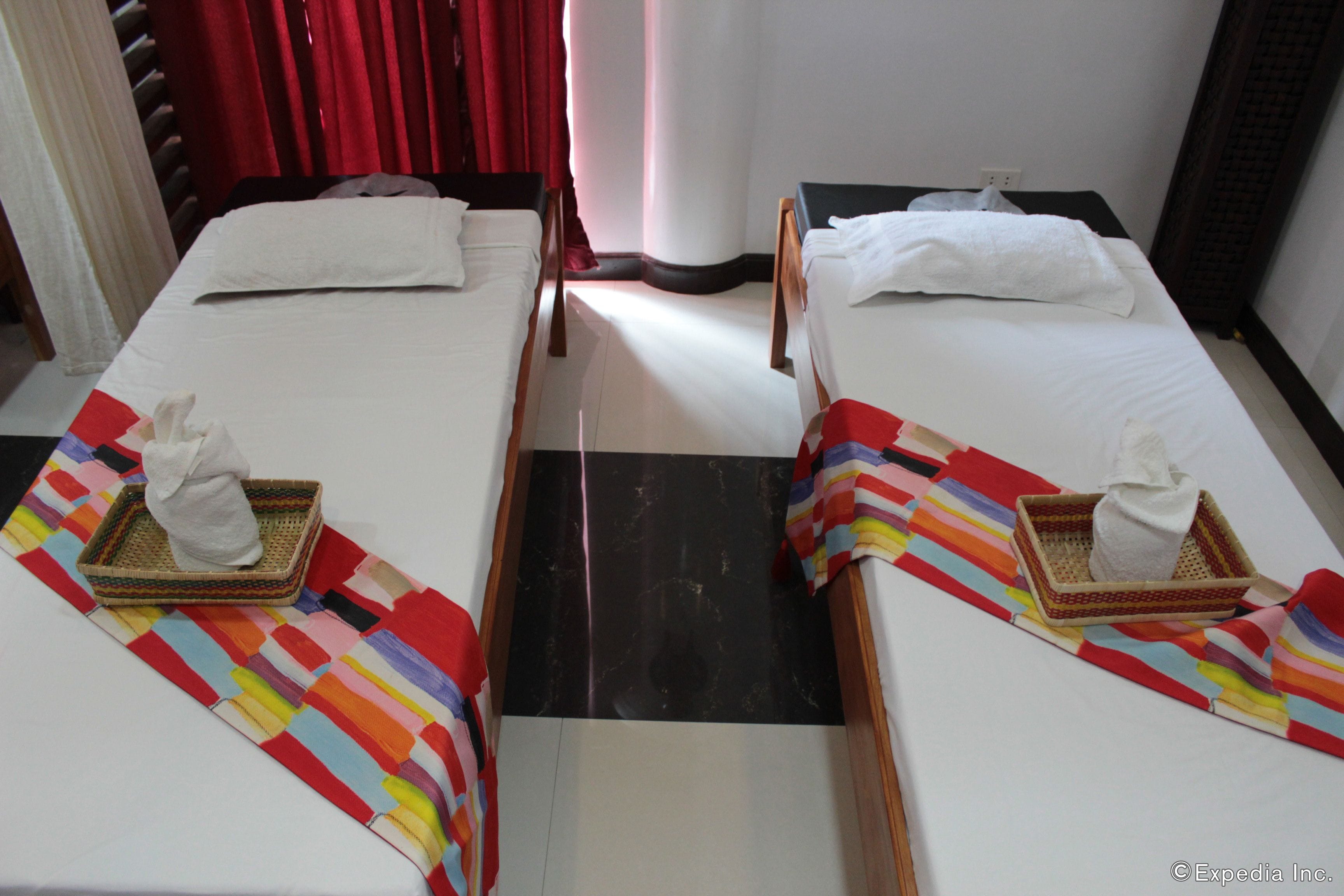 A hotel room with two single beds, each with a white pillow and a small basket containing a white towel. The room has a striped rug and a black and white floor mat.