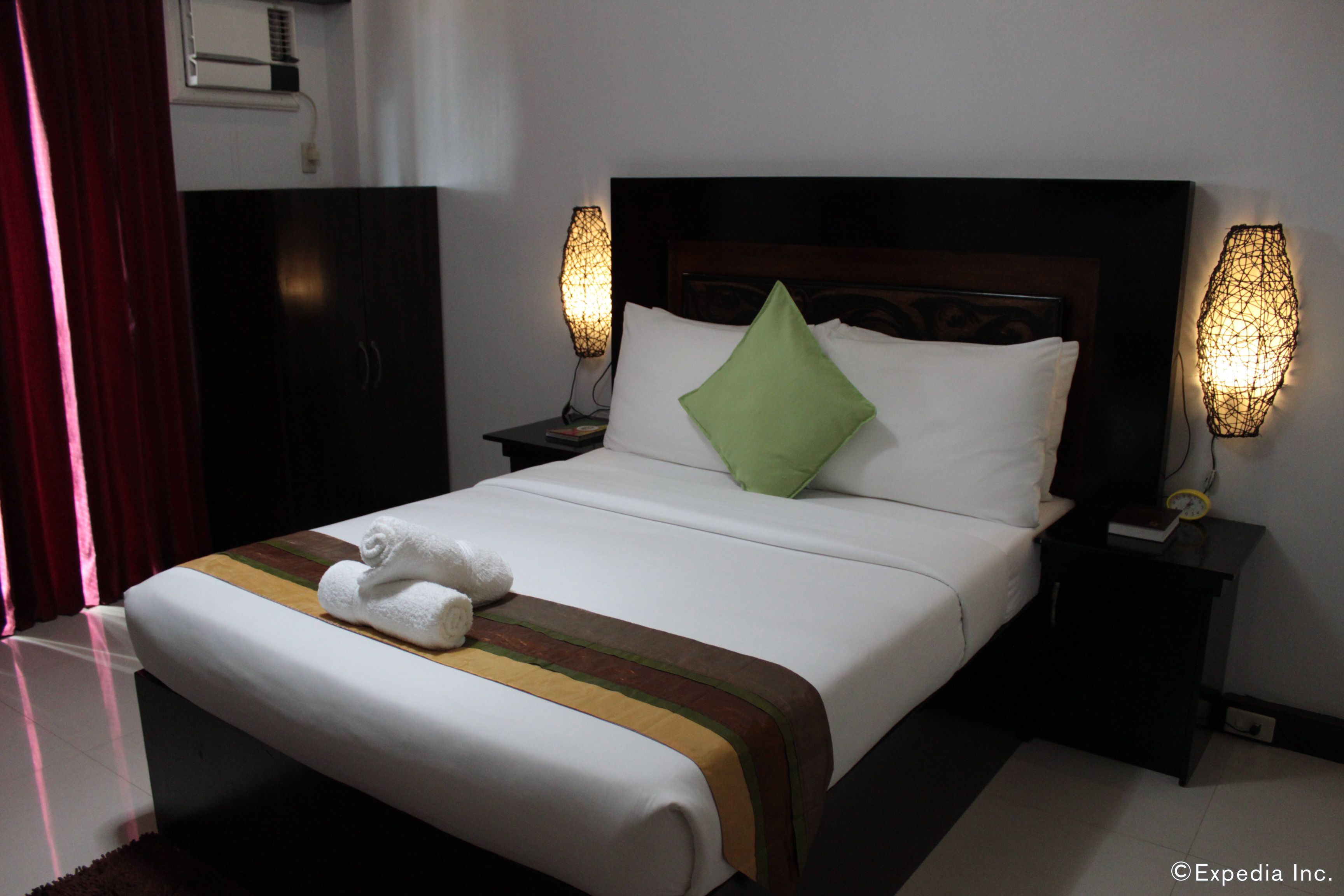 A neatly made bed with a green pillow, white linens, and a striped bedspread. There are two bedside tables with lamps, a dark headboard, and a wardrobe in the background.
