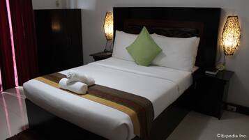 Una cama bien tendida con una almohada verde, sábanas blancas y una colcha a rayas. Hay dos mesitas de noche con lámparas, un cabecero oscuro y un armario al fondo.
