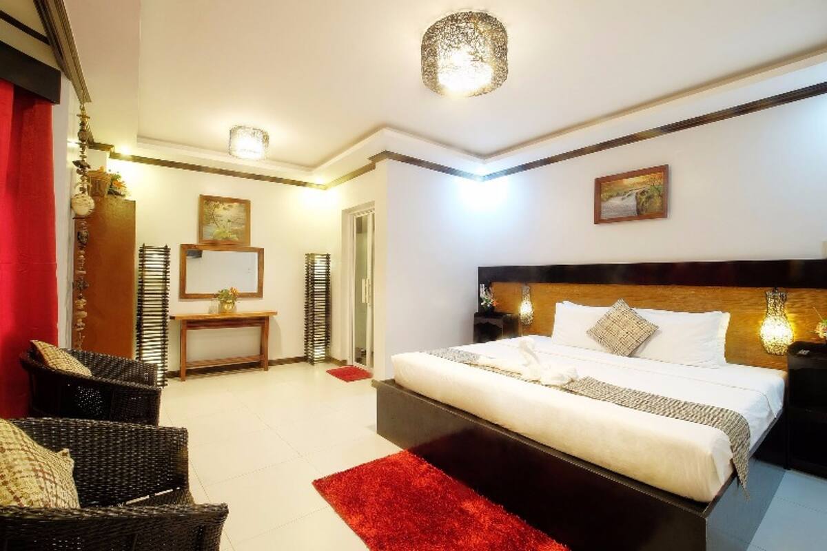 A hotel room with a large bed, a red rug, wicker chairs, a bedside table with a lamp, and a painting on the wall.