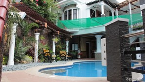 Un área de piscina con una piscina curva, rodeada por un patio con sillas y plantas. Hay varios niveles de balcones con barandillas y un paseo cubierto.