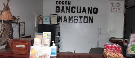 A reception desk with a sign reading "CORON BANCUANG MANSION," a wooden tip box, and various informational brochures.