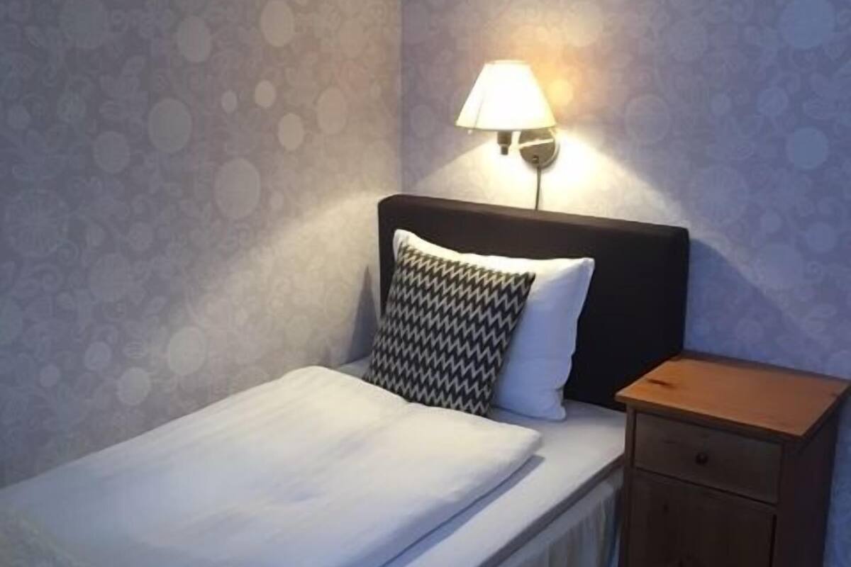 A neatly made bed with a white comforter and a patterned pillow, a bedside table with a lamp, and a wall with a circular pattern.