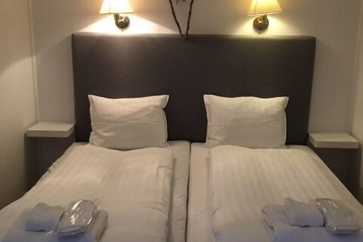 A hotel room with two beds, each with a pillow and a folded towel. There are two bedside lamps on either side of the headboard.
