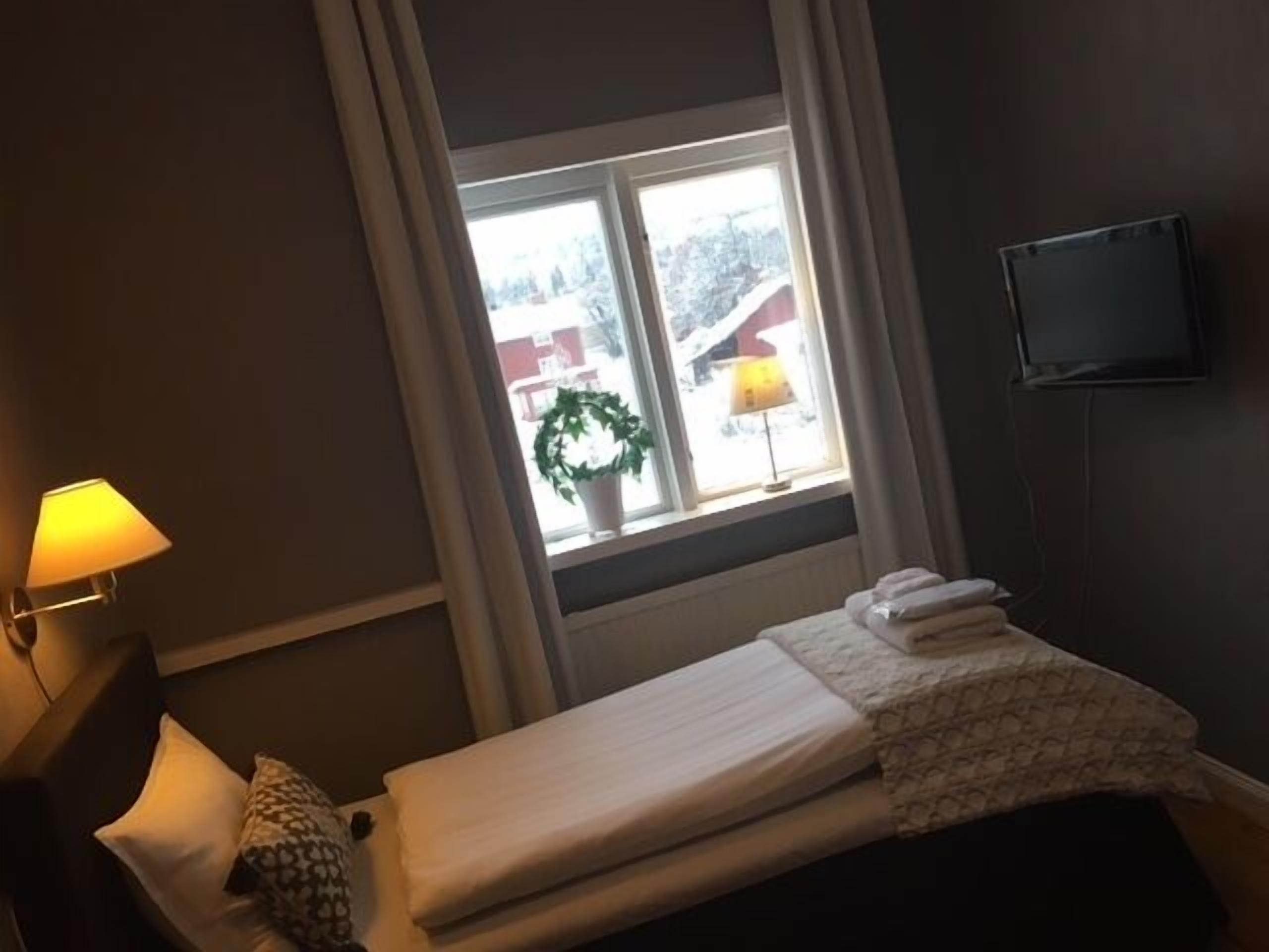 A hotel room with a bed, a window with curtains, a lamp, and a television on the wall.