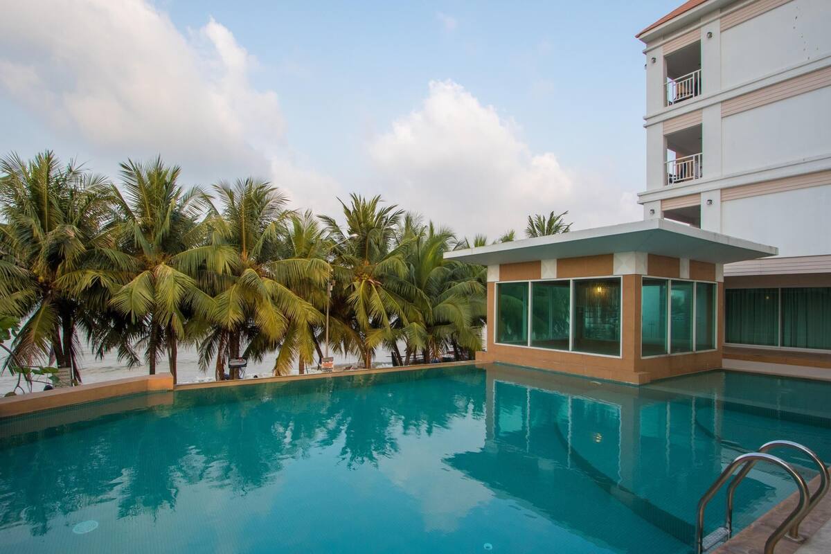 Uma piscina com água azul cristalina, rodeada por palmeiras e um edifício moderno com grandes janelas.