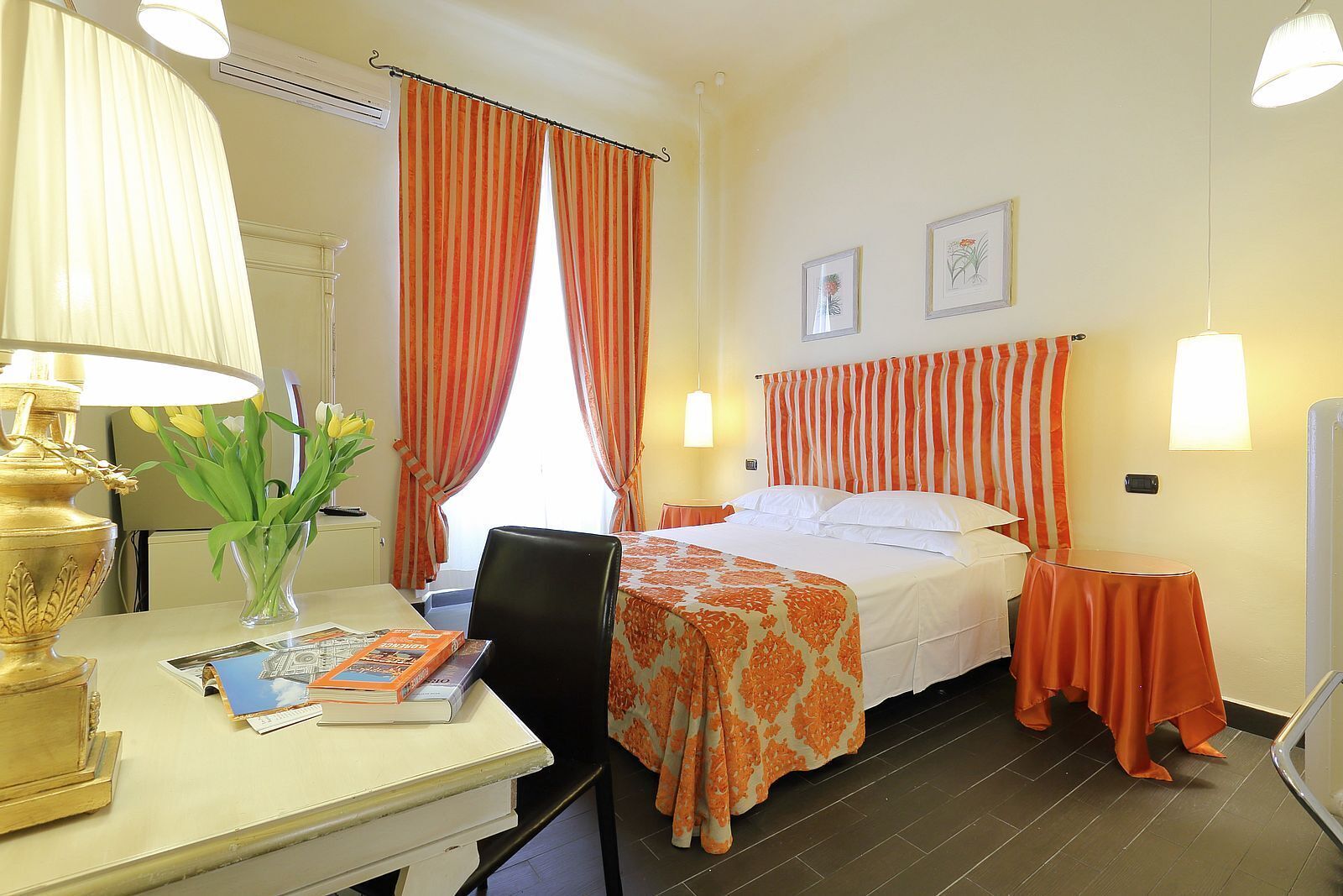 A hotel room with a bed, a desk with a lamp, a vase with flowers, and a chair.