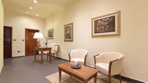 Un hall de hotel con un mostrador de recepción de madera, dos sillones a rayas, una mesita con lámpara y cuadros con motivos florales en las paredes.