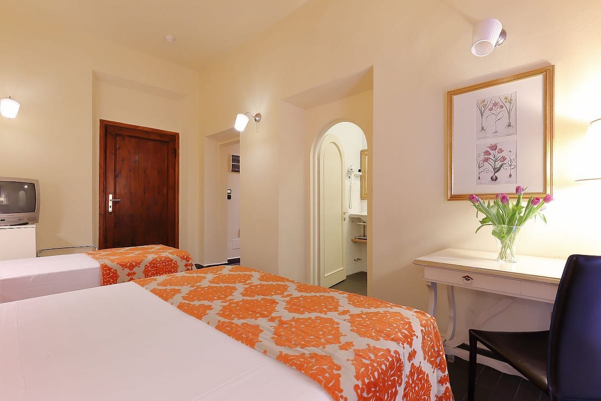 A hotel room with two beds, a desk with a chair, a television, and a framed botanical illustration on the wall.