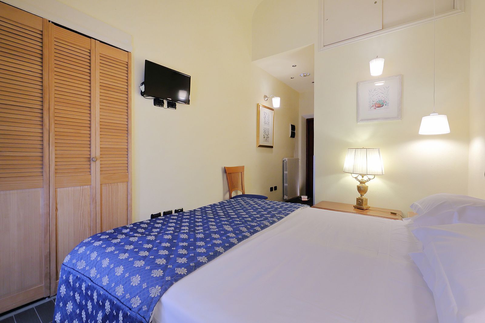 Habitación de hotel con cama, televisor montado en la pared, lámpara de noche y un cuadro enmarcado.