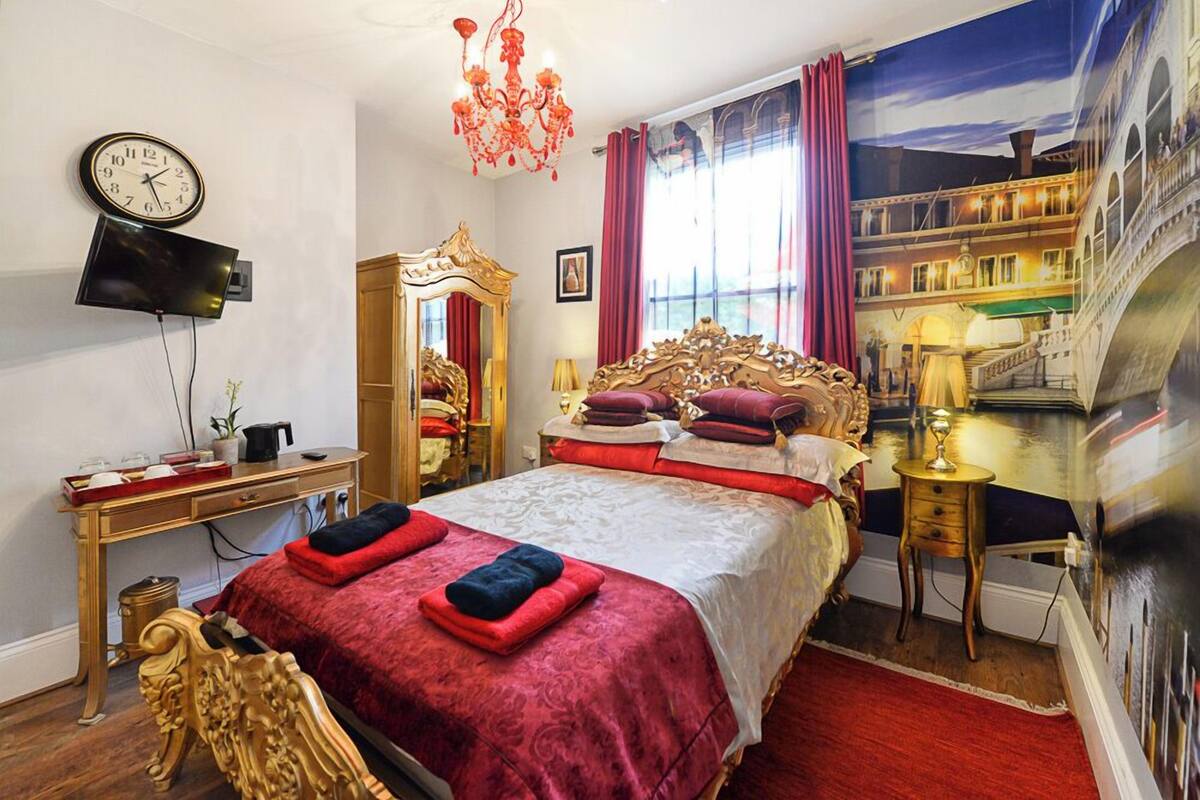 A bedroom with a large bed, a television mounted on the wall, a chandelier, and a painting of a bridge.