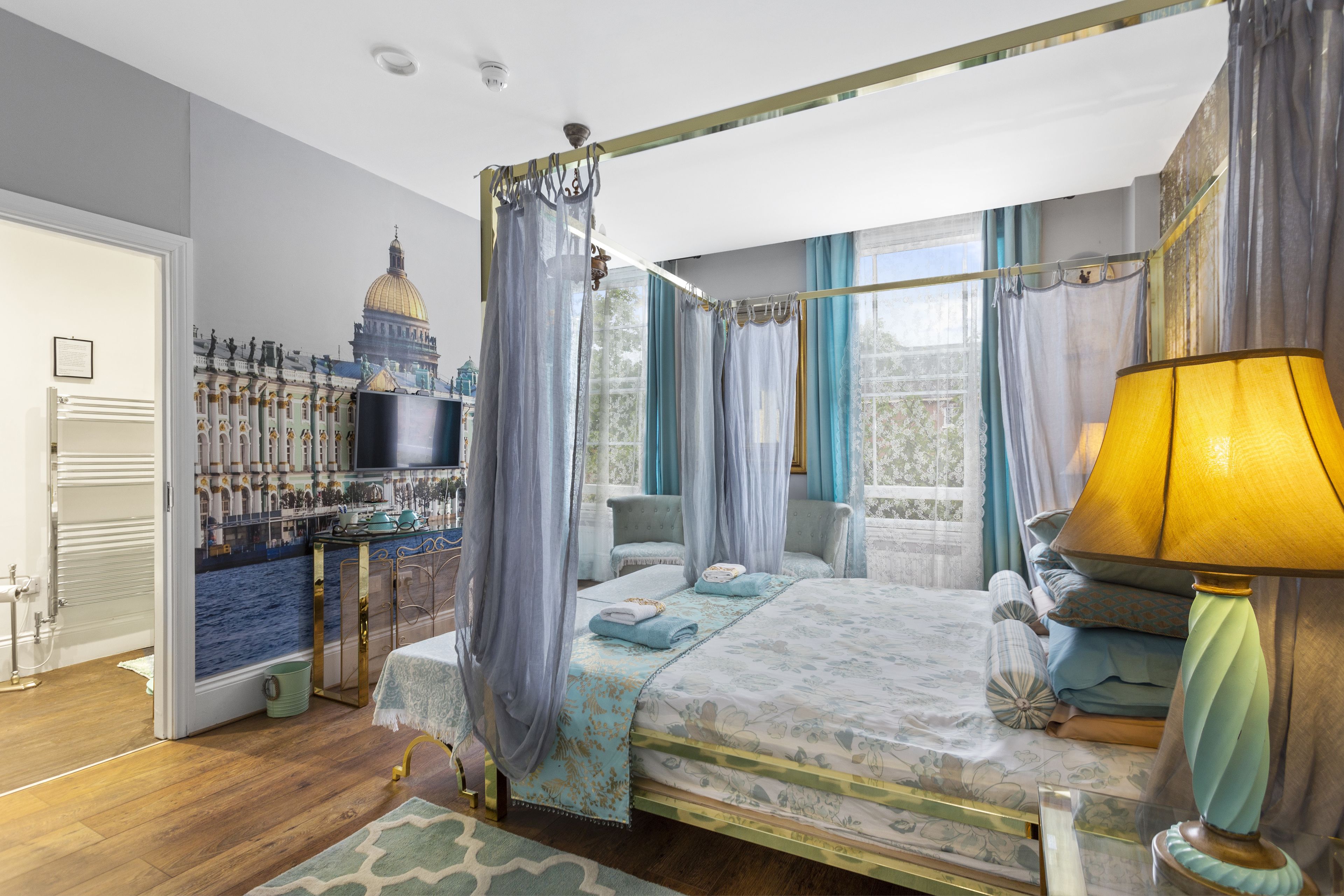 A four-poster bed with a canopy, a television, a lamp, and a view of a building through the window.
