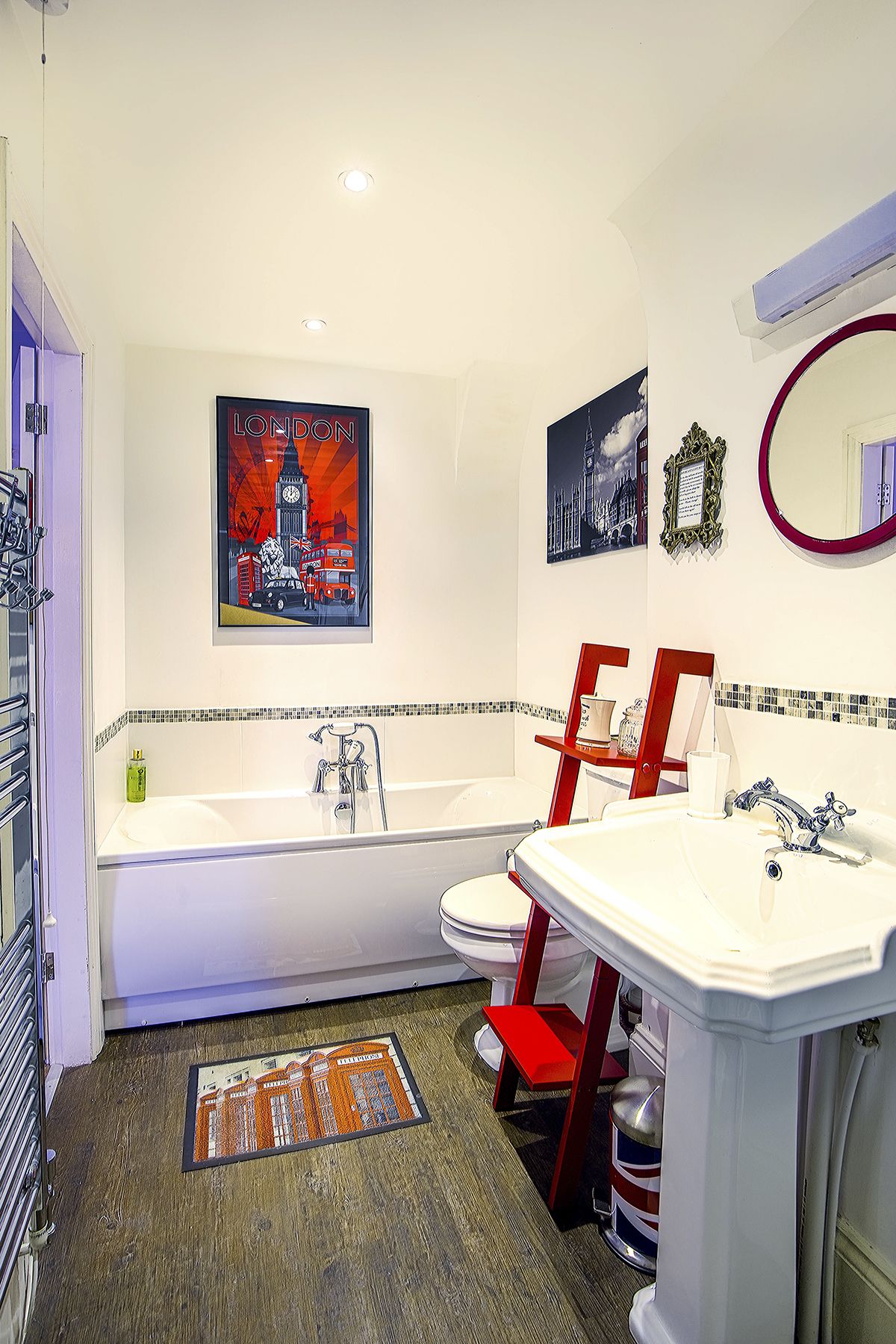A modern bathroom with a bathtub, toilet, and sink. There are two framed pictures on the wall, a mirror, and a red chair.