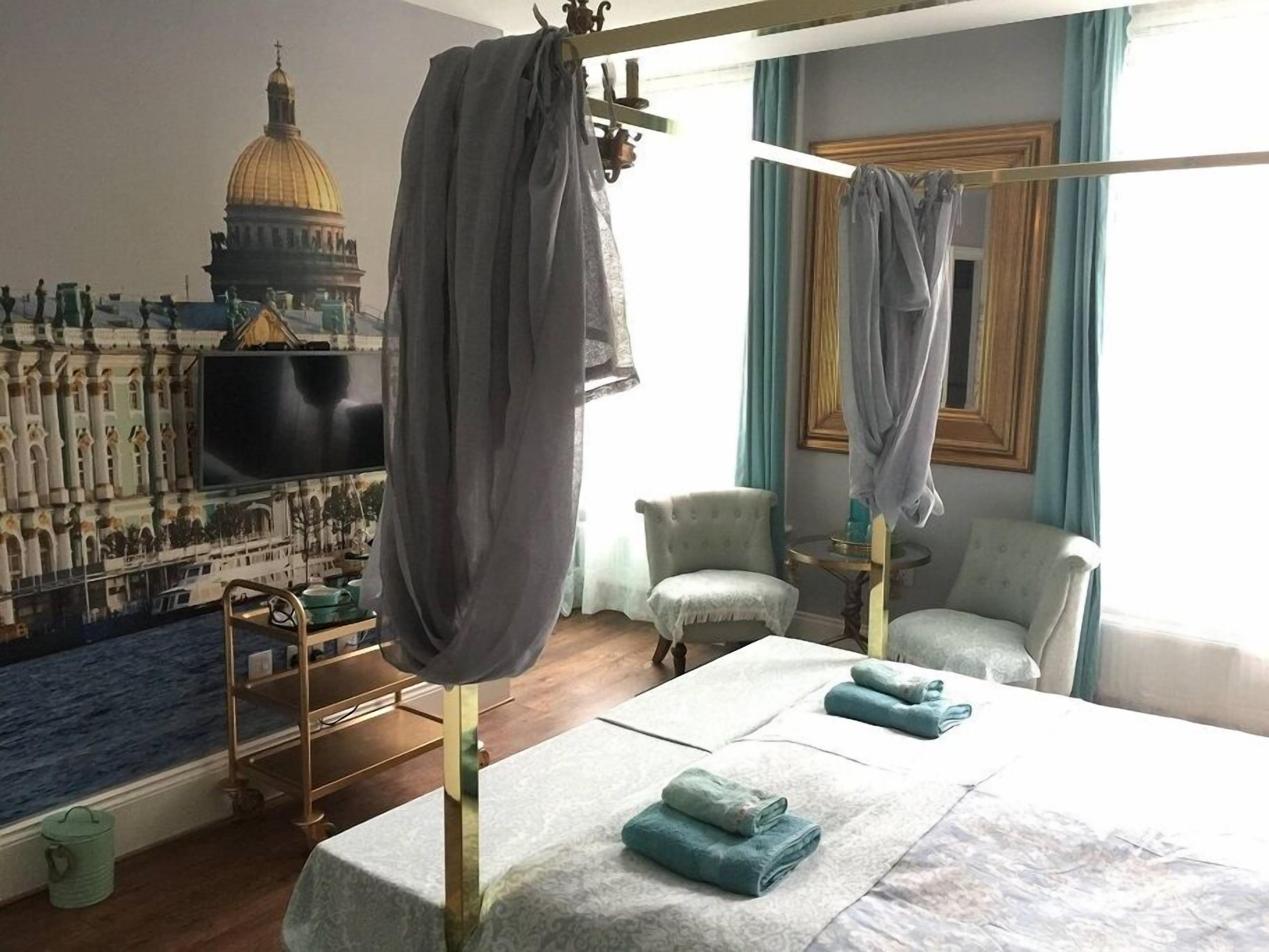 Quarto de hotel com um grande mural de uma paisagem urbana, uma cama com toalhas dobradas com cuidado, uma mesa redonda pequena com uma luminária e uma cadeira.