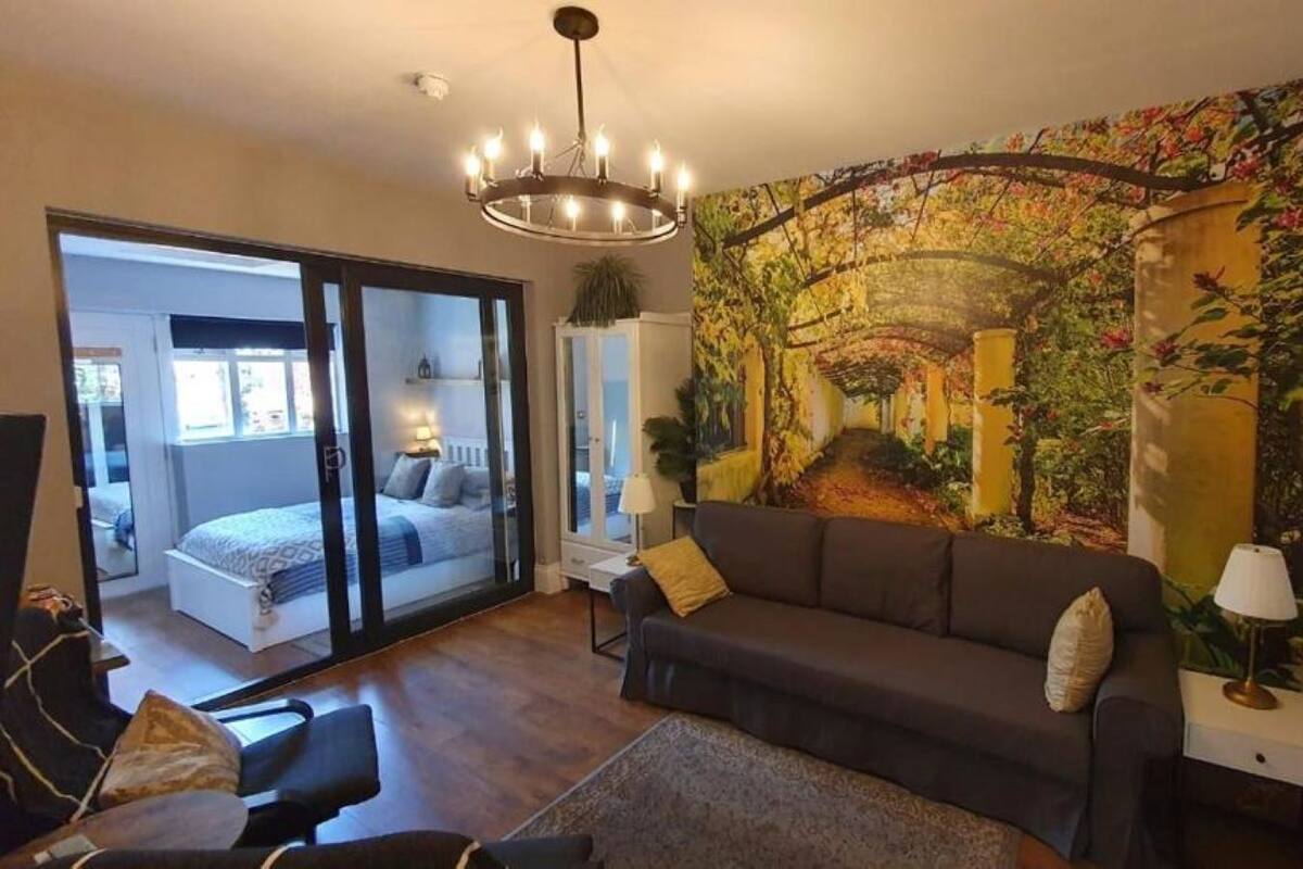 A living room with a sofa, a coffee table, and a large painting on the wall. There is a sliding glass door leading to a bedroom with a bed and a window.