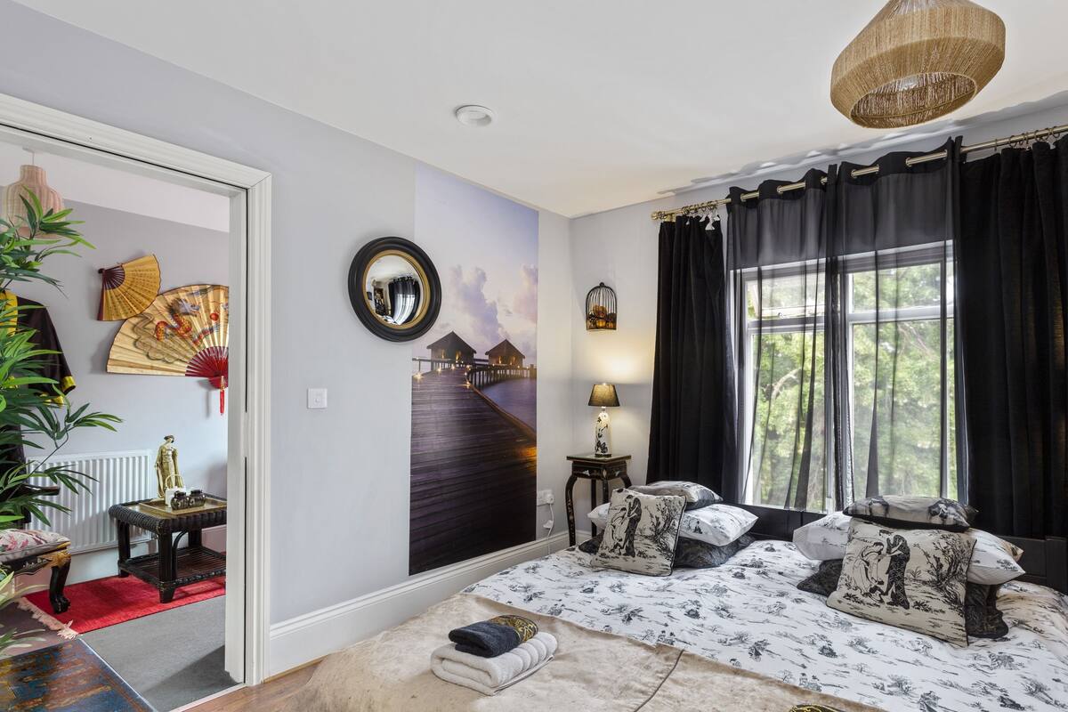 A room with a bed, a mirror, a lamp, a window with curtains, a wall with a fan, and a red rug.