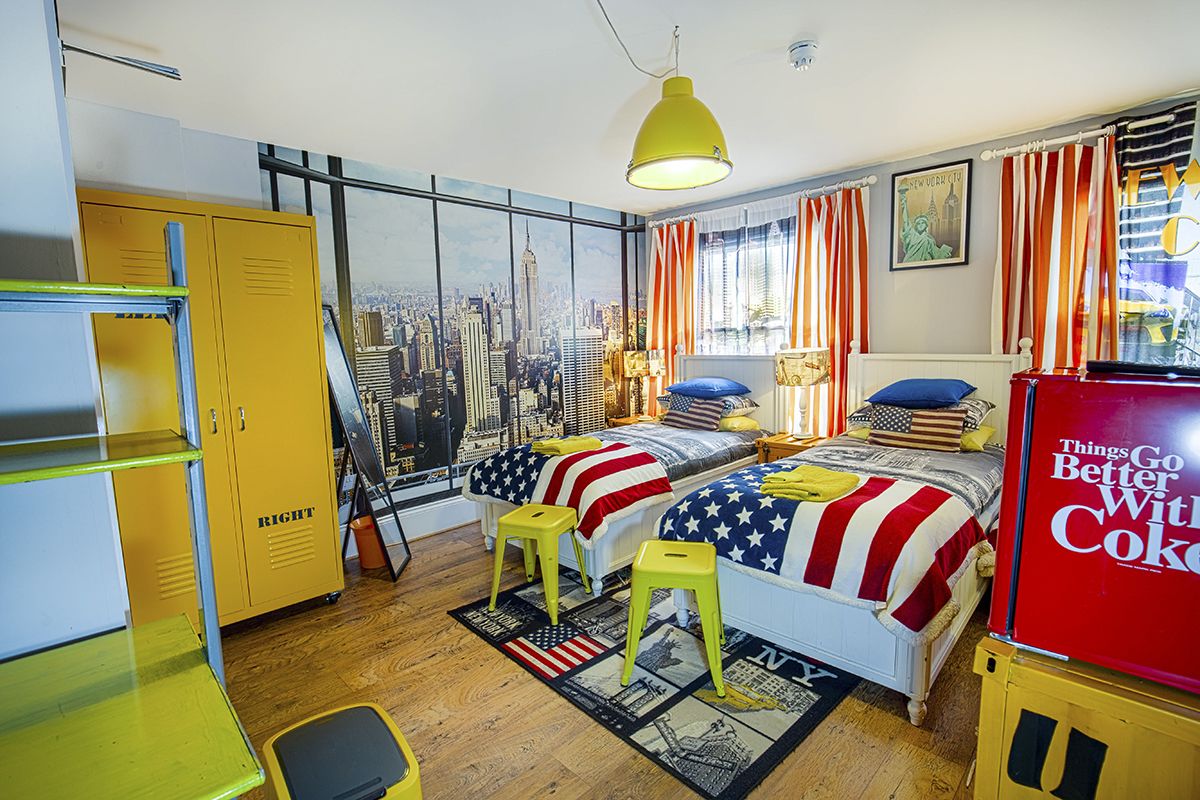 A bedroom with two beds, a cityscape view, yellow lockers, and a red refrigerator.