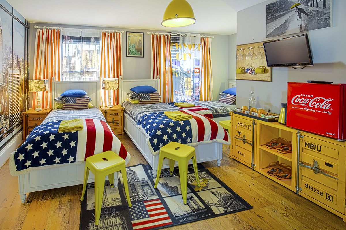A hotel room with two beds, each adorned with an American flag blanket, a television, a Coca-Cola refrigerator, and a shoe cabinet.