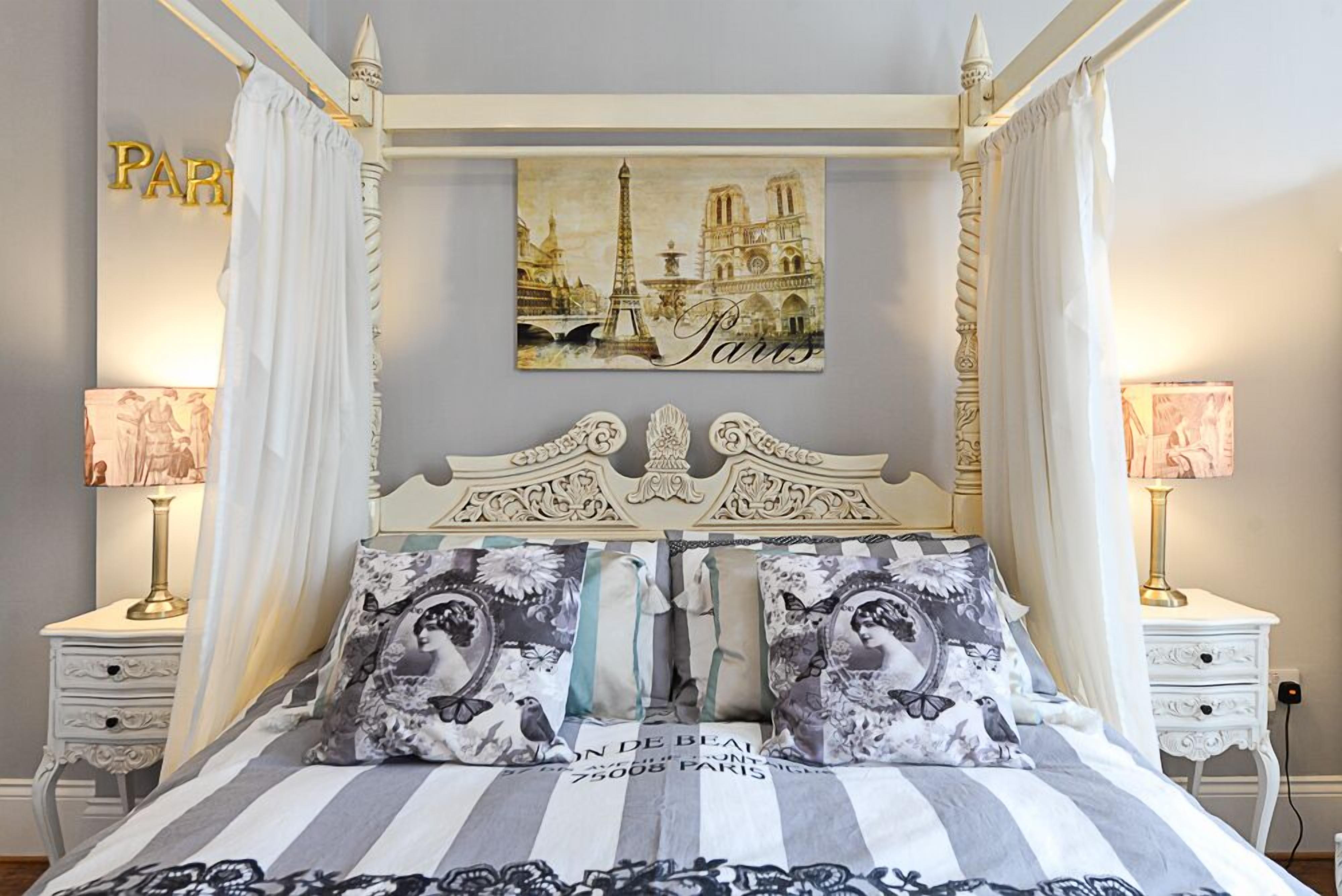A four-poster bed with a striped bedspread and decorative pillows, a white headboard, and a Paris-themed wall art.