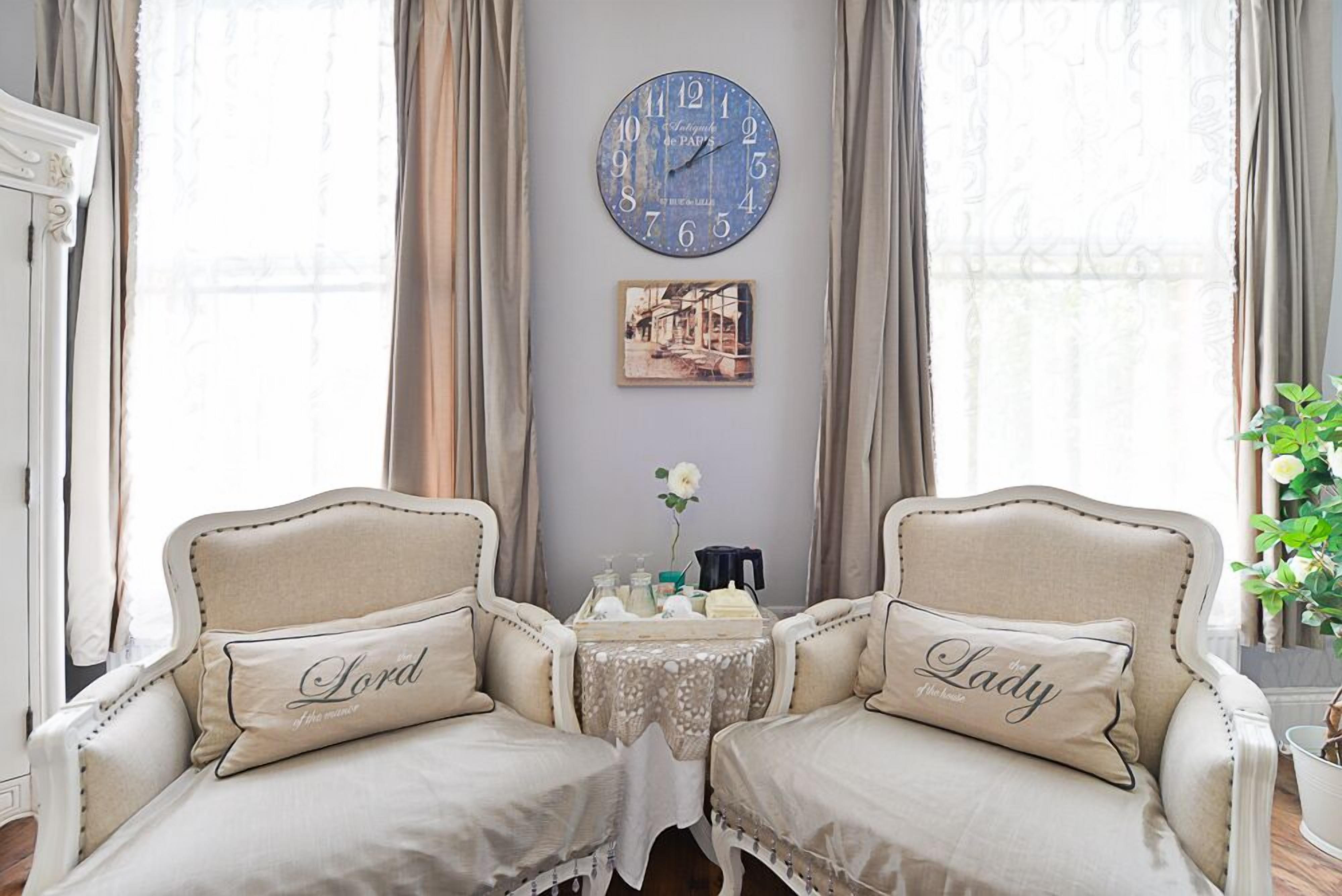 A room with two armchairs labeled "Lord" and "Lady," a round wall clock, a small table with a tray, and a framed picture on the wall.