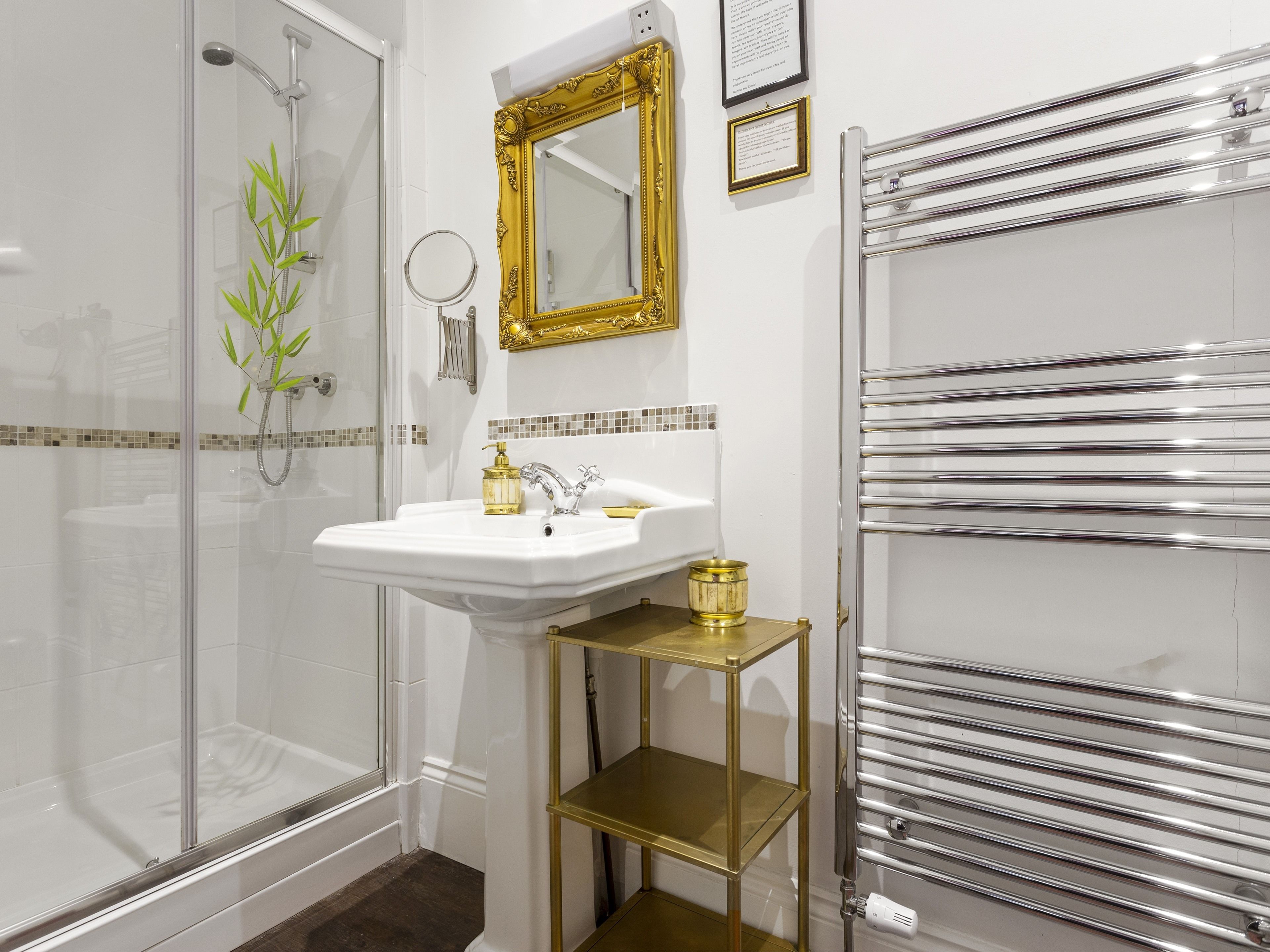 A bathroom with a glass shower enclosure, a white sink with gold fixtures, a gold-framed mirror, and a chrome towel rack.