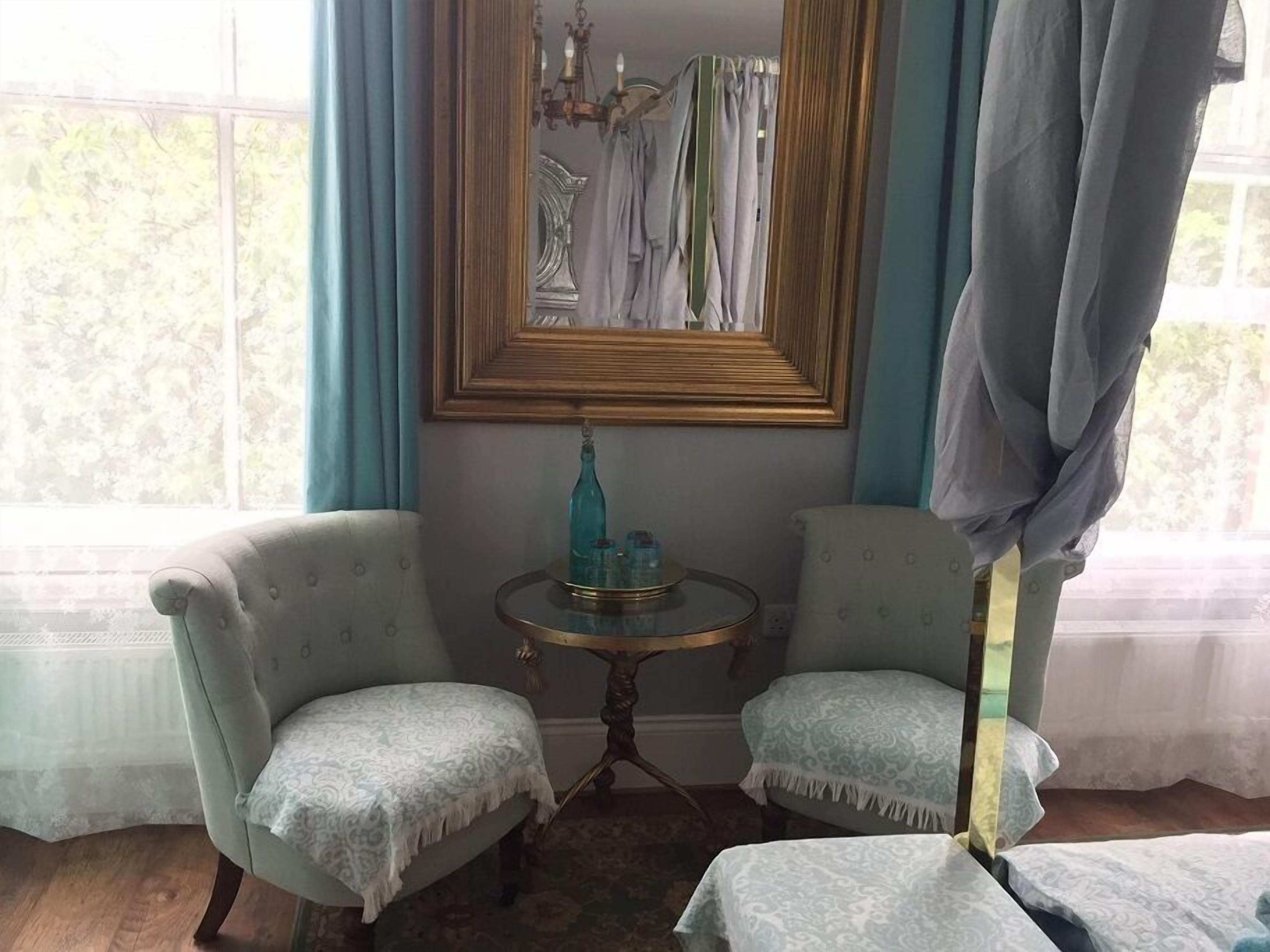 Quarto com duas poltronas estofadas, uma mesinha redonda com uma garrafa e um espelho acima.