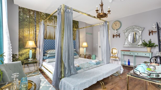 A four-poster canopy bed with a gold frame, two bedside lamps, a mirror, and a small table with decorative items. - Mama's Inn Boutique Guest House (Nottingham)