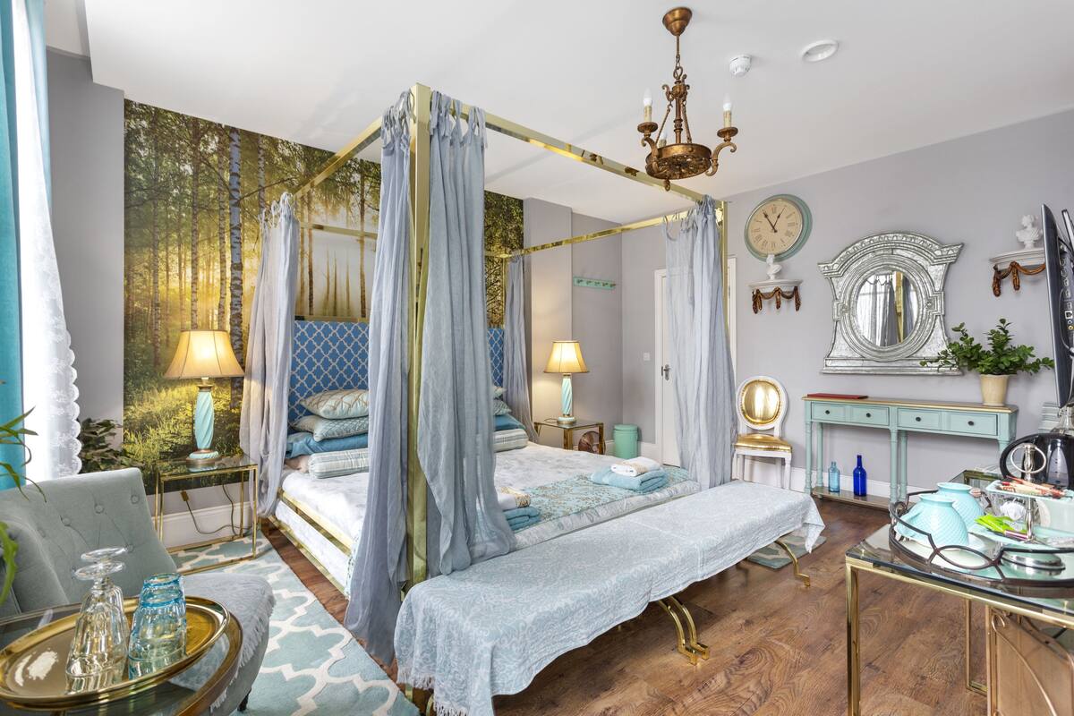A four-poster canopy bed with a gold frame, two bedside lamps, a mirror, and a small table with decorative items.