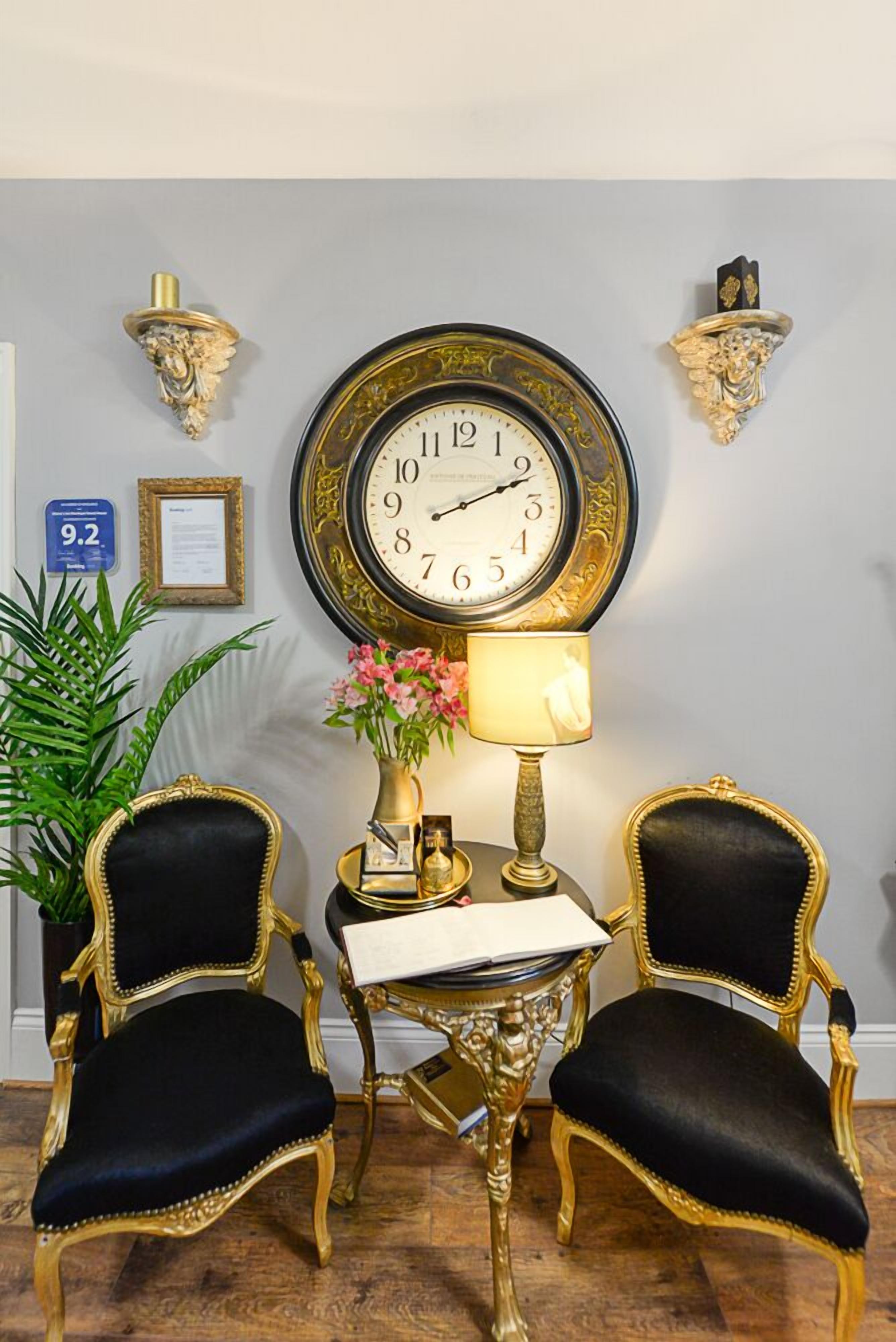 A room with two black upholstered chairs with gold trim, a round wall clock, a small table with a lamp, a vase with flowers, and a framed certificate on the wall.