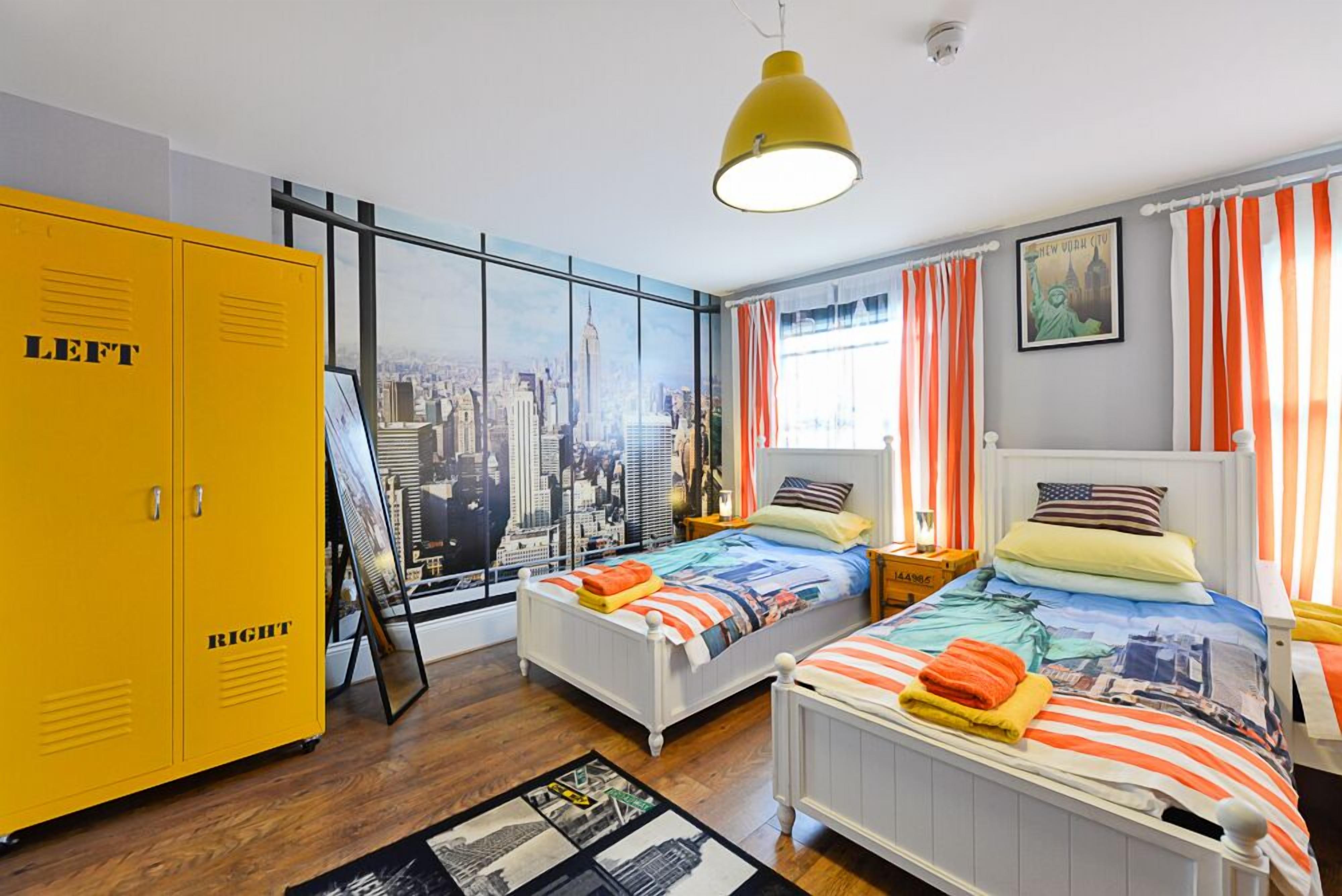 Une chambre avec deux lits, une armoire jaune, une fresque représentant un paysage urbain et une grande fenêtre avec des rideaux orange.