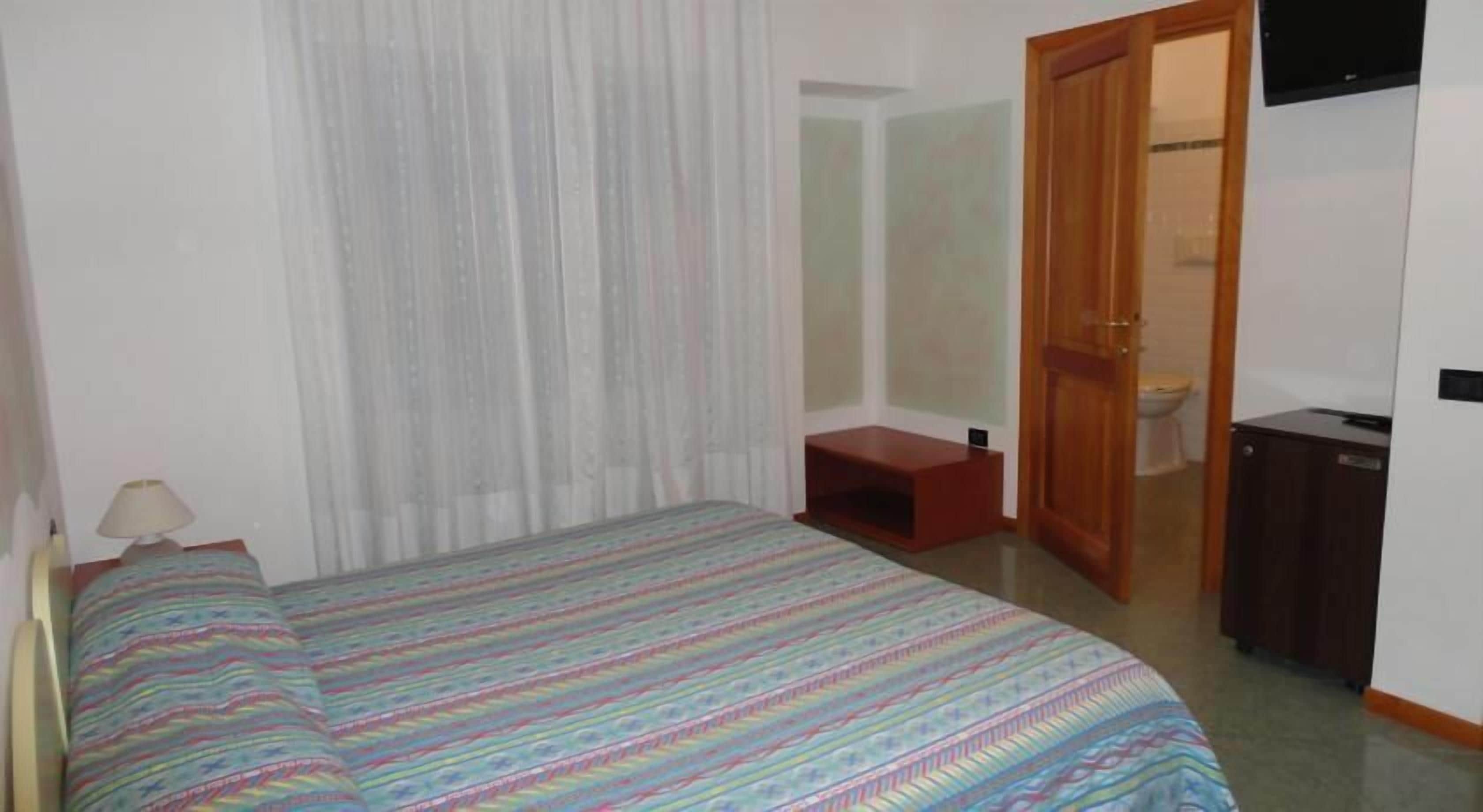 A bedroom with a bed, a bedside table, a lamp, a small wooden stool, a television, and a door leading to a bathroom.