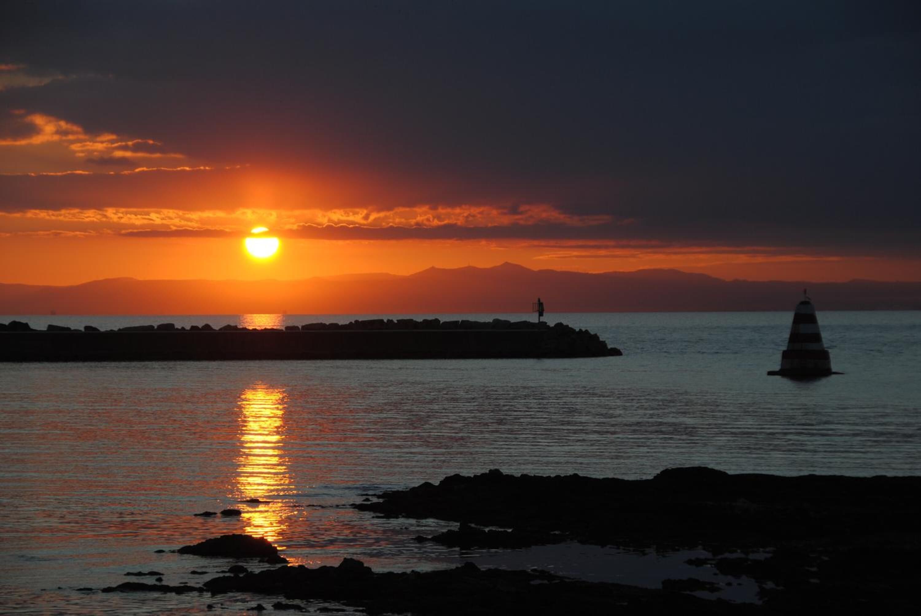 A sunset over a body of water with a lighthouse and a striped buoy.