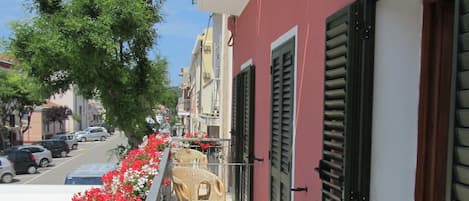 A balcony with wicker chairs and red flowers, a red building with shutters, and a street view.
