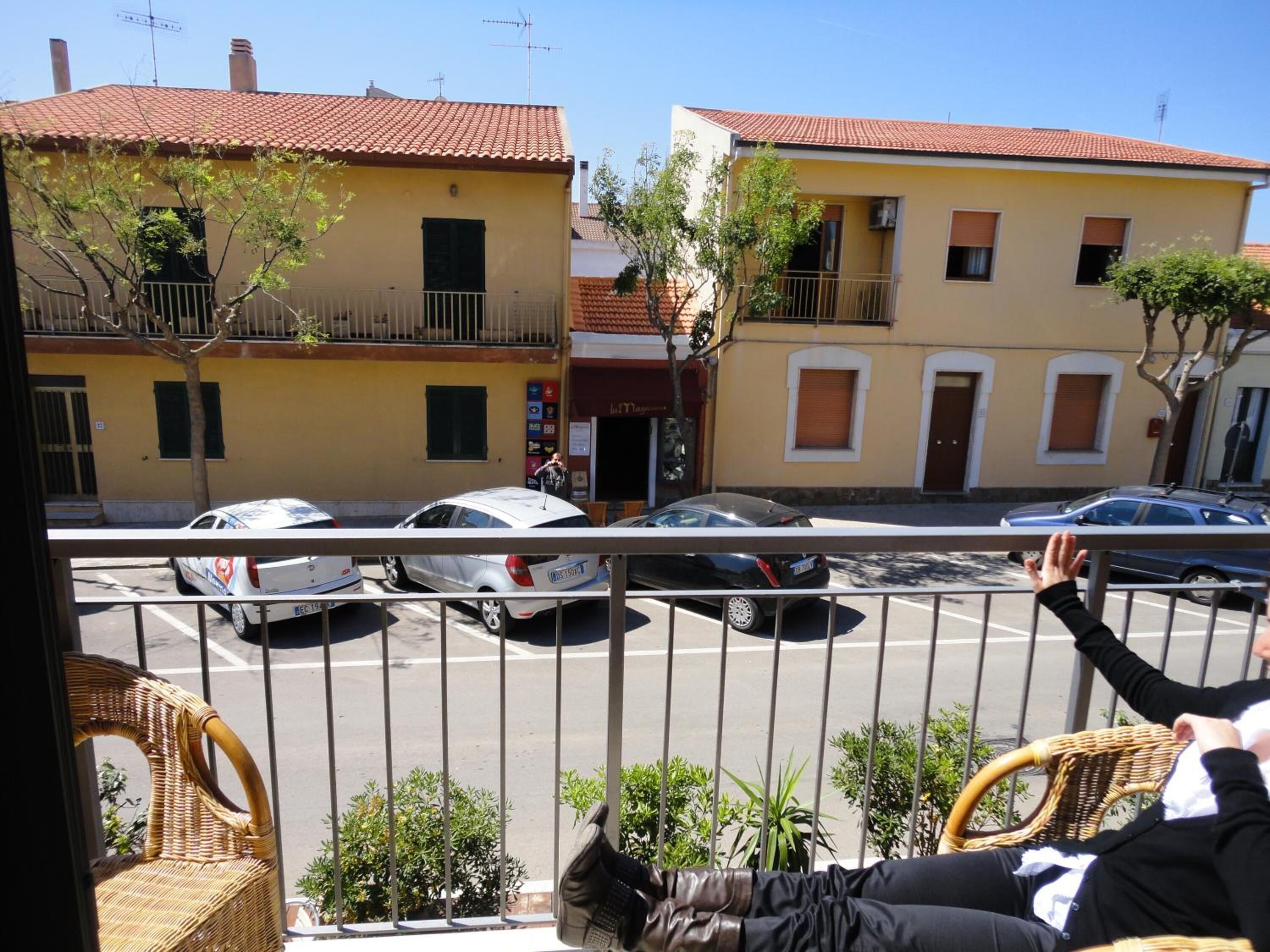 Un balcon avec des fauteuils en osier et une vue sur une rue bordée de voitures garées et d’immeubles.
