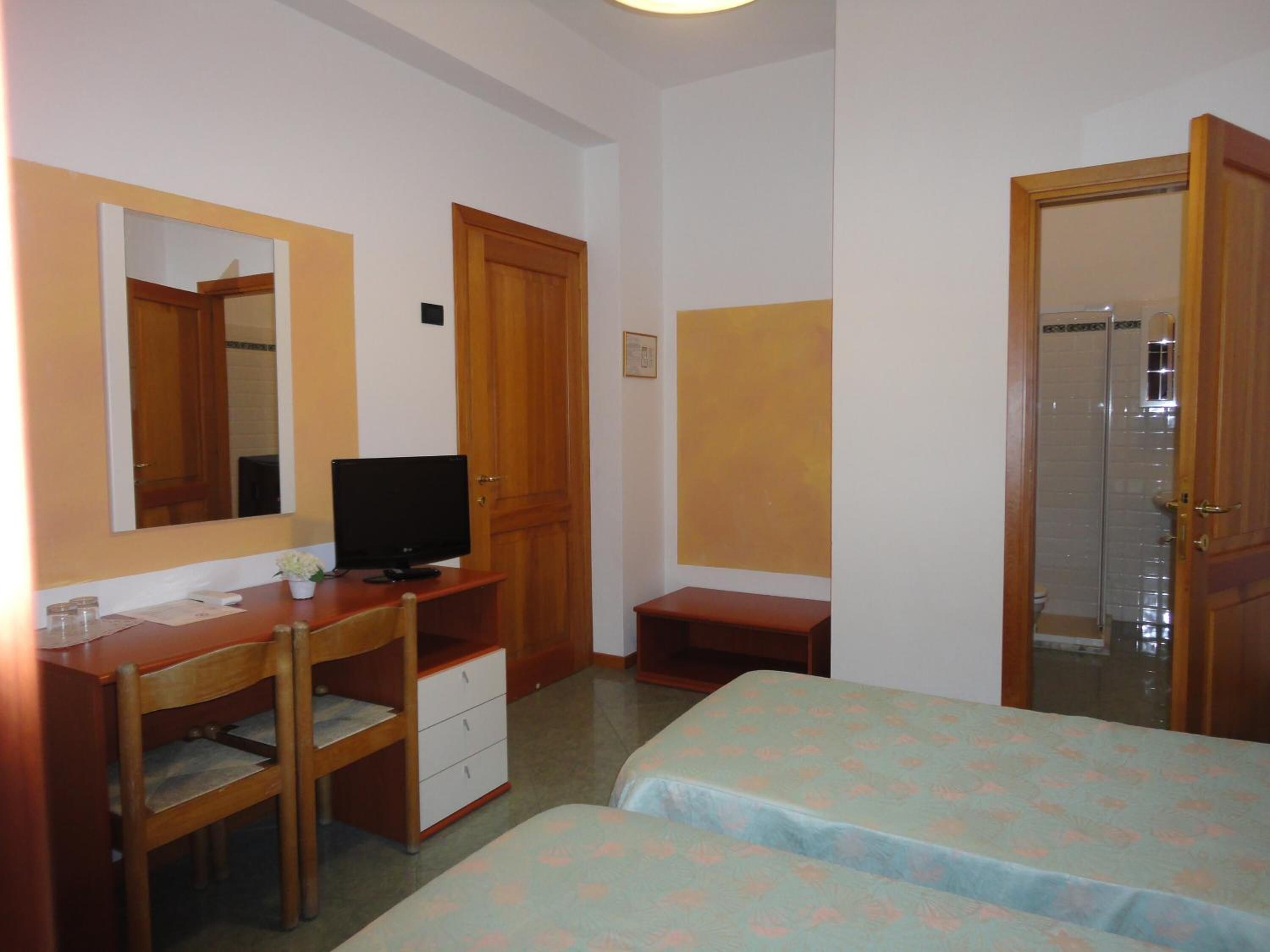Ein Hotelzimmer mit einem Holztisch, einem Fernseher, zwei Betten mit floralen Überwürfen, einem Spiegel und einer Tür zu einem Badezimmer.