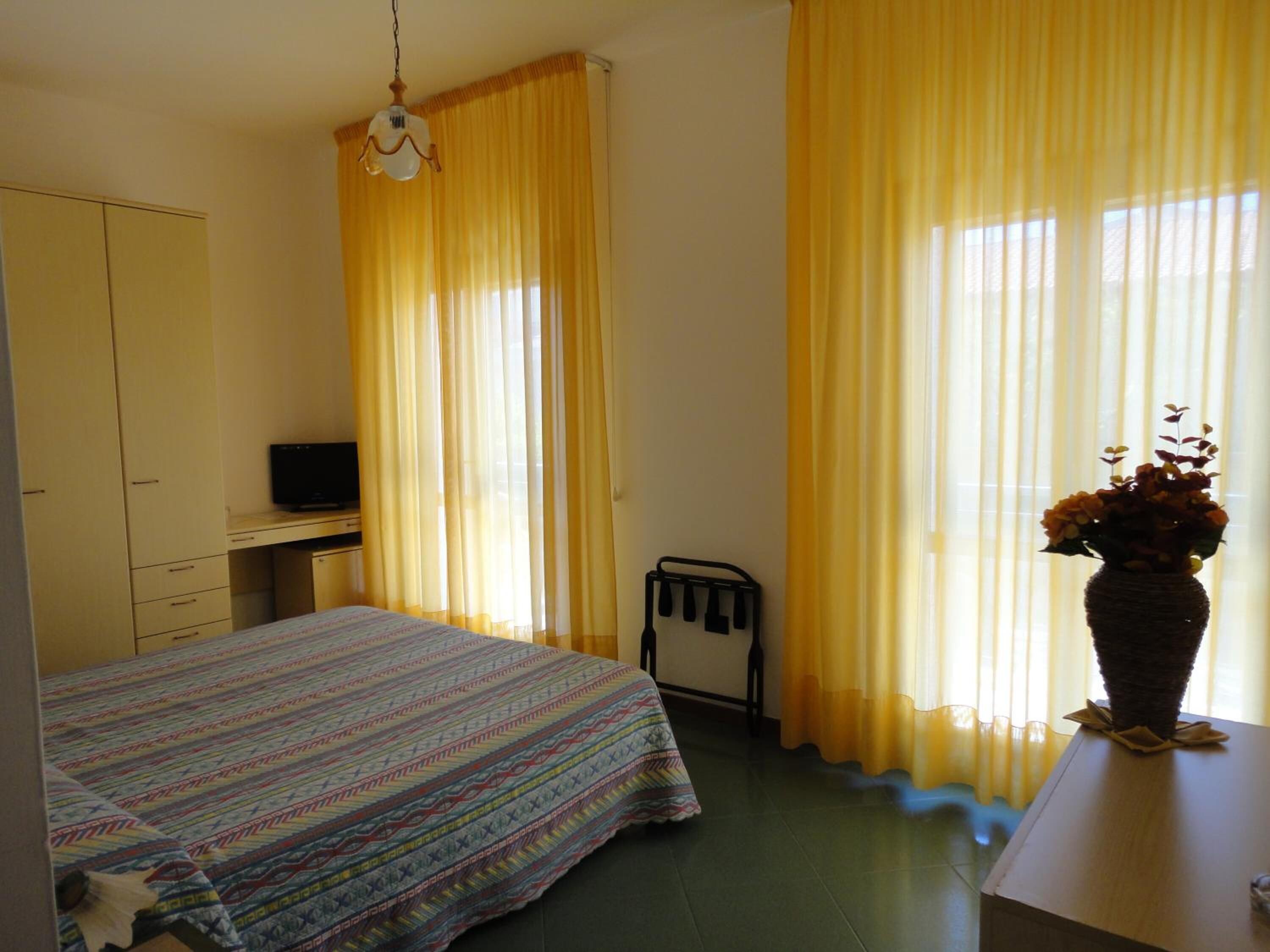A bedroom with a bed, a desk, a chair, a television, and a window with yellow curtains.