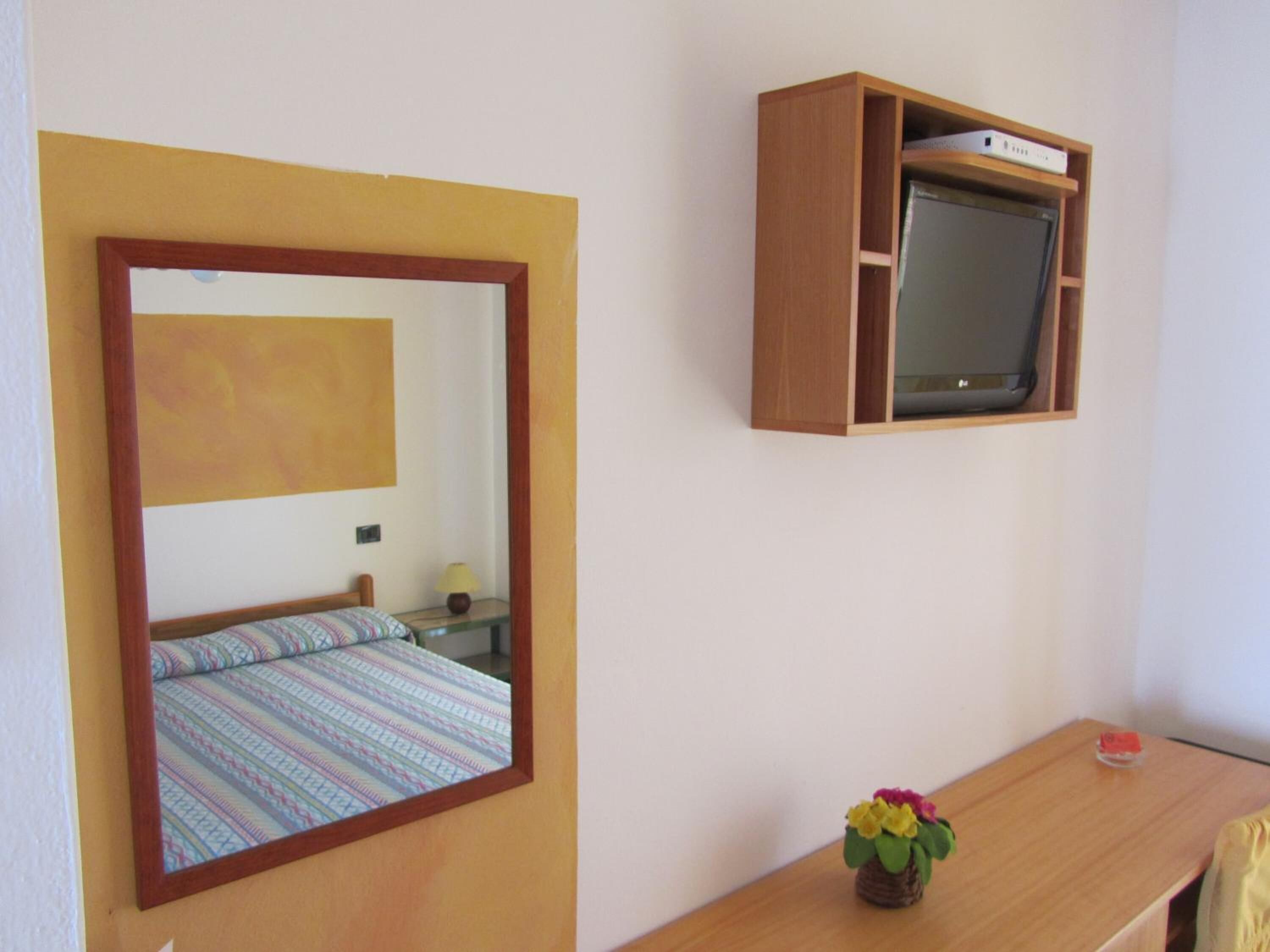 A bedroom with a bed, a bedside table with a lamp, a small table with a flower, a TV mounted on the wall, and a mirror.