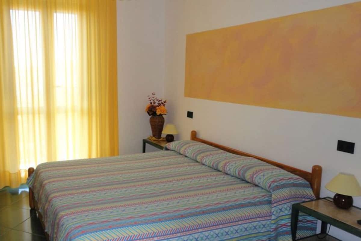 A bedroom with a bed, curtains, a nightstand, a lamp, and a painting on the wall.