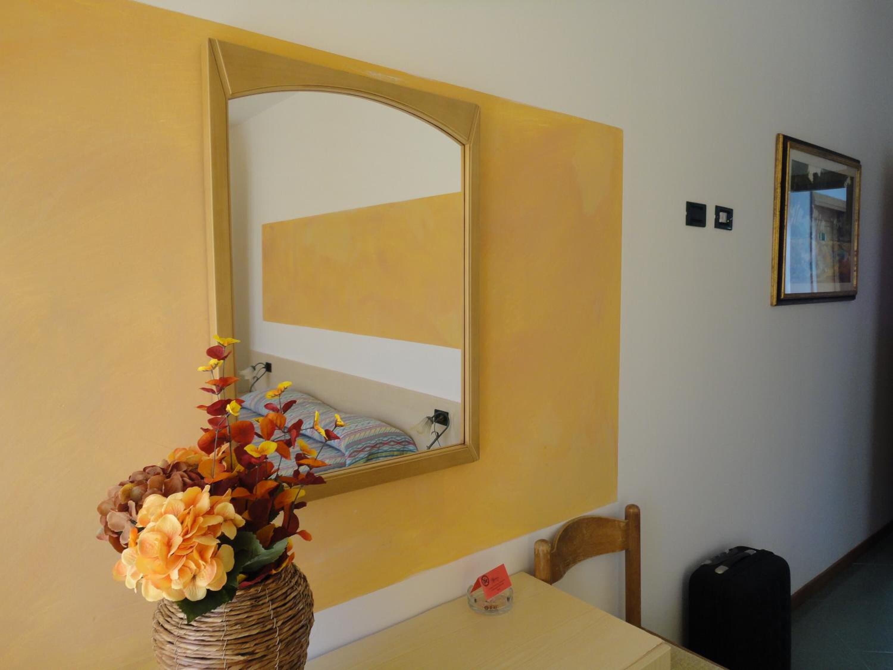 Una habitació amb una paret groga, un mirall, una gerra de vimet amb flors, una cadira, una taula i un quadre emmarcat.