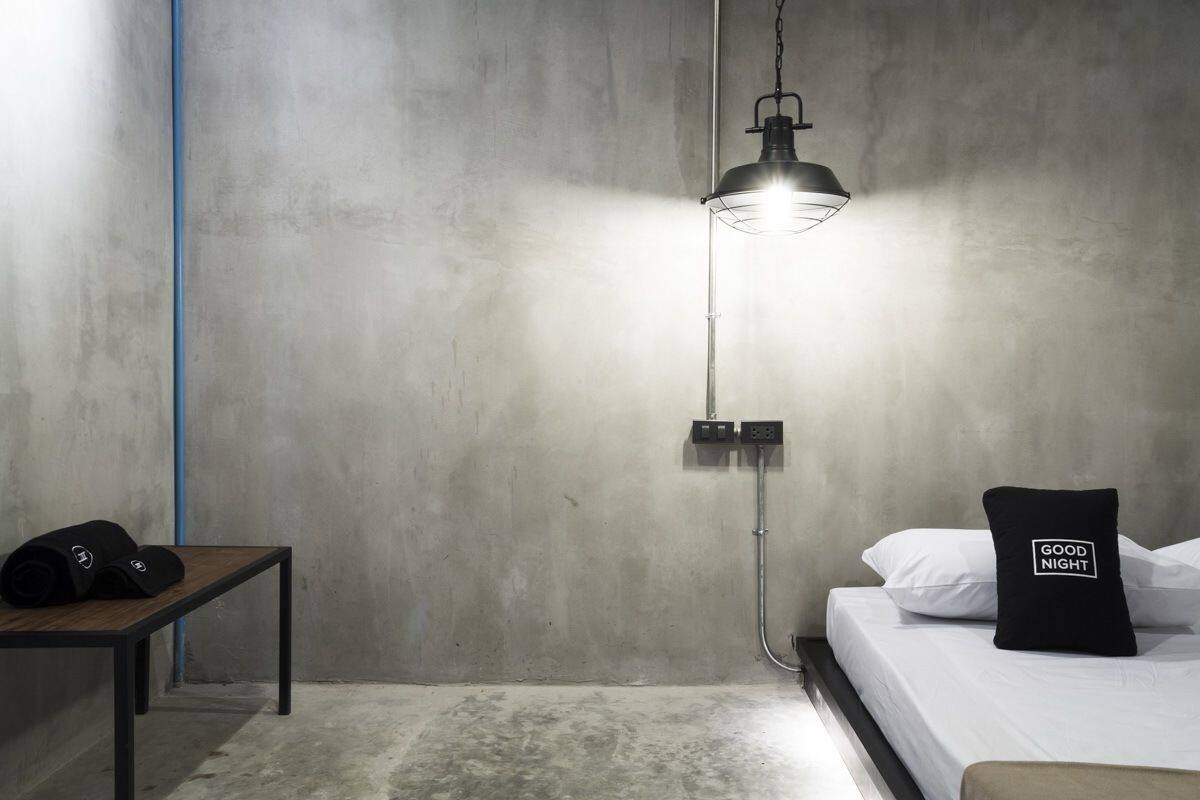 A bedroom with a bed, a bedside table with a black object, a hanging lamp, and a wall-mounted power outlet.