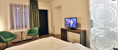 A hotel room with a bed, two green chairs, a TV on a stand, a door with a circular design, and a window with sheer curtains.