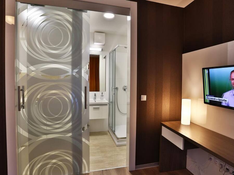 Une chambre d’hôtel moderne dotée d’une porte en verre donnant sur la salle de bain, d’un téléviseur à écran plat fixé au mur et d’une table de chevet avec une lampe.