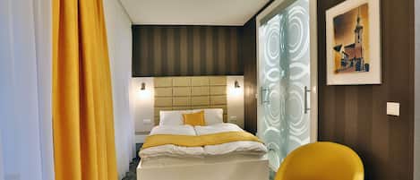 A hotel room with a bed, a yellow chair, a painting on the wall, and a door with a glass panel.